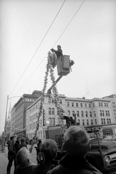 City workers install holiday decorations and lights on light poles on the corner of South Carroll and W. Main Streets at the Capitol Square. A man is seen in a small crane cab by a telephone line as children below look up at him.  