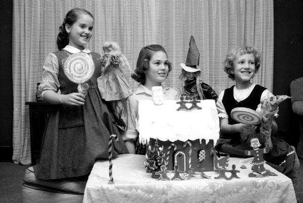 Three young friends are shown with puppets and setting ready to give a performance of Hansel and Gretel. Of their earnings, they intend to give $3 to the Empty Stocking Club. They are Barbara Torkelson (daughter of Mr. and Mrs. William Torkelson), Janet Ficken (daughter of Mr. and Mrs. Robert Ficken), and Nan Wegner (daughter of Mr. and Mrs. Robert Wegner). Mrs. Wegner has given many puppet performances on TV, and is the advisor to her daughter and friends. The girls are seen performing behind a gingerbread house and holding large lollipops with their puppets.