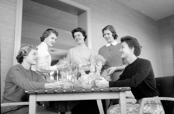 Some of the young people working on committees for the Maple Bluff Counry Club's holiday party for the high school are shown. From left to right are: Sydney Fish, Mary Ann Hopkins, Ann Whitford, Linda Elliott, and Ann Holford. There are decorations on the table in front of them.