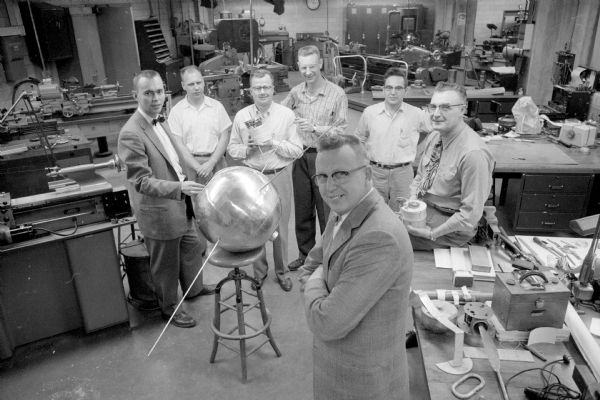 Image shows seven men in a U.W. research lab posing with a life-sized model of a satellite. Professor Verner Suomi of the meteorology department led a team that constructed instruments that were to be included inside one of the U.S. Vanguard satellites. Suomi hoped to study the flow of heat from the Sun to the Earth and then back to space. The seven men are (left to right in the back): Professor Wayne Swift, Andrew Grondahl, Harry Miller, Charles R. Stearns (also of meteorology), William Hauser and E J. Romare. In the foreground is Professor Suomi, team leader. All men except Stearns and Suomi were with the College of Engineering.