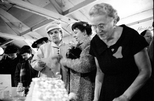 Mrs. William Reiter (left) and Mrs. Conrad Jaeger (center), both of McFarland, looking at a recipe while Mrs. Carl Edmonds browses through the food items at the Christmas tea for Dane County Homemakers clubs. Mrs. Edmonds has a pin in the shape of a bell on her dress.