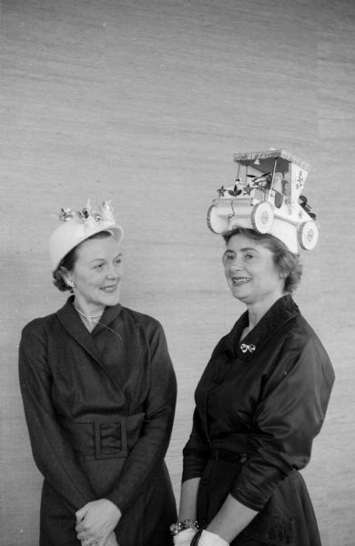 Elsa Johnson looks up at a hat topped with a large wheeled carriage worn by Betty Berst. Johnson was style show chairman for 1957. Berst was the Maple Bluff Country Club's social chairman in 1958.
