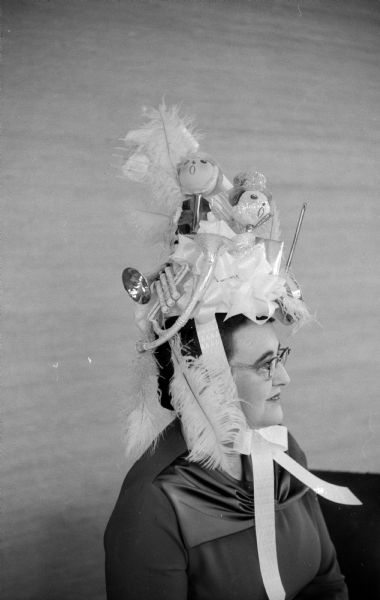 Eleanor Nerdrum, program chairman for the Maple Bluff women's group in 1956-57, is wearing a hat richly decorated with a variety of Christmas tree ornaments like feathers, bows and small musical instruments that is tied under her chin with large ribbon to hold it up.