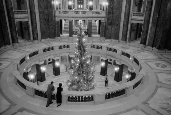 A view from above of the tall decorated 34-foot balsam tree in the Capitol Building rotunda. The tree was grown in Marquette County and purchased from the Coyne Brothers Christmas Tree Co. for $45. Elevated view from a balcony down onto the opening of the second-floor, from where the top of the tree with tinsel and lights stands. A man and a woman are standing at the balcony in the foreground.
