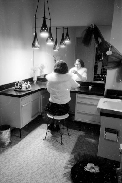 The tri-level home of I. Franklin and Arline Houseman, 821 Lakewood Boulevard in Maple Bluff, was redecorated with her choosing the color scheme. The "strictly for women" powder room featured pink walls, fixtures, and built-in vanity, accented with black counter-tops and hanging lamps. She is sitting on a stool in front of the mirror at the vanity.