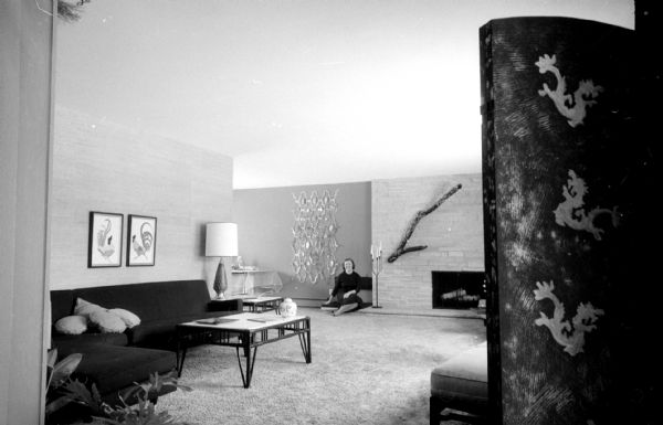 The tri-level home of I. Franklin and Arline Houseman, 821 Lakewood Boulevard in Maple Bluff, was redecorated with her choosing the color scheme. The living room shows contemporary structural and furnishings design with gray walls and carpeting. The room is also decorated with art on the walls and an Asian screen with Chinese dragons. Mrs. Housman is sitting on the left side of the fireplace.