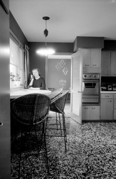 The tri-level home of I. Franklin and Arline Houseman, 821 Lakewood Boulevard in Maple Bluff, was redecorated with her choosing the color scheme. The kitchen features a free-form breakfast bar with purple walls and driftwood finish cabinets. The oven console can be seen built into the cabinetry. Mrs. Houseman is sitting at the kitchen table by the window and talking on the phone.