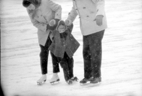 A young girl trying out a new pair of skates with the help of her parents at the Tenney Park lagoon. It is snowing.
