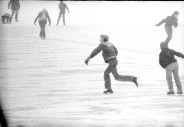 An experienced ska ter isstriding across the ice at the Tenney Park lagoon. Other people are skating, and it is snowing.