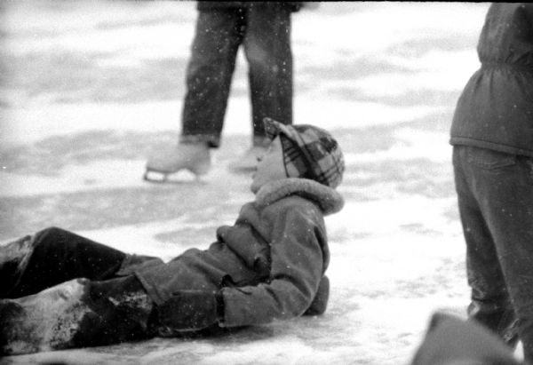 A young skater has fallen on the ice at the Tenney Park lagoon. It is snowing.