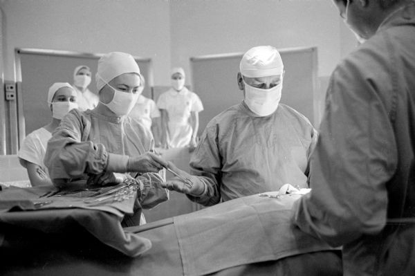 Student nurse, Mary Jo Oxem (left) in surgical garb assisting a surgeon in an operating room. As one of the 125 students of St. Mary's School of Nursing, she is posing for a series of photographs taken during a typical day of a senior student nurse.