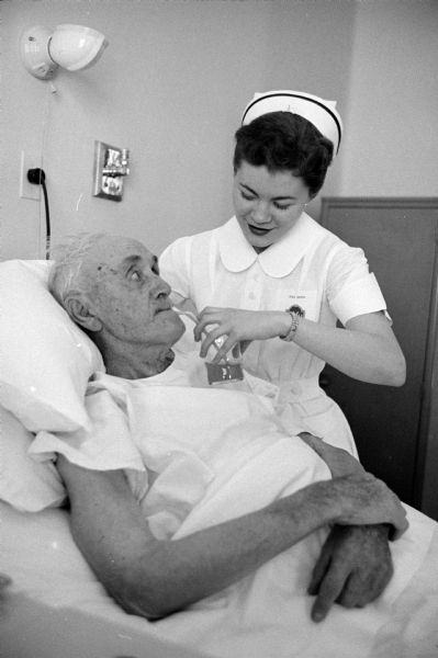 Student nurse, Mary Jo Oxem, giving water to post surgical patient, Ambrose Bronner. As one of the 125 students of St. Mary's School of Nursing, she is posing for a series of photographs taken during a typical day of a senior student nurse.   

