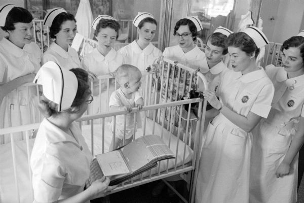 Student nurse, Mary Jo Oxem, with seven of her classmates at the bedside of a young child. They are listening to their nursing instructor, Mrs. James Sapietal. 

Student nurses are (left to right): Jo Anne Alex, Sheboygan; Joanne Brenshan, Beloit; Sally O'Brien, Antigo; Jeanette Johns, Cuba City; Rose Marie Caygill, Madison; Rosalie Studer, Monroe; and Mary Jo Oxem, Madison. In the foreground is Mrs. Sapietal. 

As one of the 125 students of St. Mary's School of Nursing, Mary Jo is posing for a series of photographs taken during a typical day of a senior student nurse.