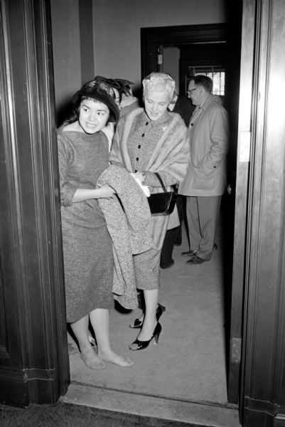 Governor Gaylord Nelson's niece, Maya Lee (of Crestwood), is shown resting her feet as the Governor's sister, Mrs. Spencer Yates (of Roseburg, Oregon), is looking on. She has removed her shoes and is seen looking through a doorway in the Capitol.