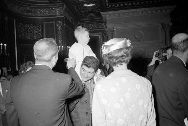 Sixteen-month old Dave Watts, son of Mr. and Mrs. Jim Watts, riding on his father's shoulders to greet Governor-elect Gaylord Nelson and Mrs. (Carrie) Nelson in the Governor's conference room after the inauguration in the Capitol.

A press man with a hand-held film camera is getting footage of the action of the Governor and First Lady.
