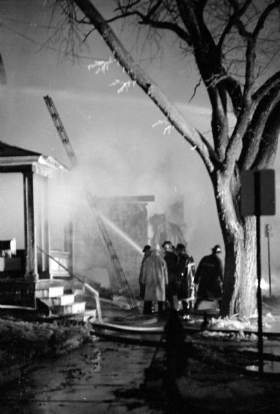 Explosion and fire in the 200 block of North Bassett Street caused the deaths of William and Gladys McGrath and destruction of five buildings. Firemen are seen training a hose on a porch in an attempt to confine the fire, which was caused by gas explosions, and a ladder is leaning against the house frame.