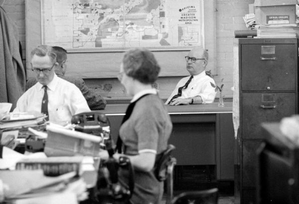 Lawrence Fitzpatrick, Managing Editor of the <i>Wisconsin State Journal</i>, sitting at his new desk speaking with someone. A woman and two men are at desks to the left in the foreground. A map of the greater Madison metropolitan area is on the wall behind Fitzpatrick.