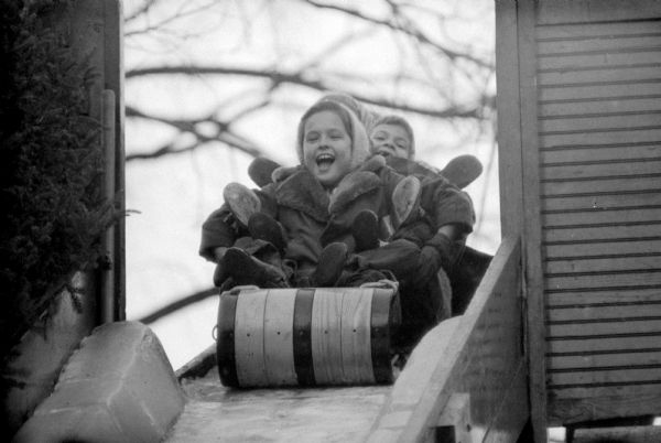 A girl is beaming in anticipation while sitting at the front of a wooden toboggan at the top of a steep slide at Olbrich Park. Another child is behind her, and by the number of shoes sticking out to the sides, there are quite a few friends about to go down the slide with her.