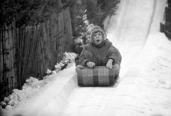 Children dressed in heavy winter clothing, on a wooden toboggan at the bottom of a steep slide erected at Olbrich Park.
