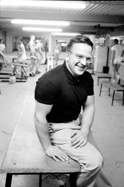 John L. "Bud" Fuller, proprietor of Madison Health Studio at 235 West Gilman street. He is sitting in a workout room, where men can be seen behind him on a treadmill, lifting weights and exercising.