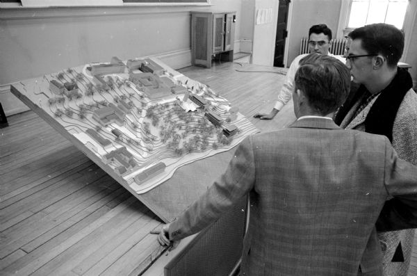 Several people looking at a topographical model of Bascom Woods and Muir Knoll by the surrounding buildings between Elizabeth Waters Hall, the Carillon Tower and the Memorial Union. They are attending a hearing about locating a new building in the area.