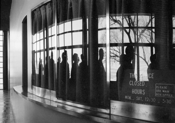 Reflections in the curved windows of the theater box office cause distortions as patrons wait in line for the office to open. The people are silhouetted against the reflection of Lake Mendota in the glass. 