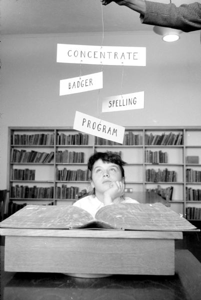 The cover picture for the Badger Spelling Bee pamphlets to be mailed by the Wisconsin State Journal to schools in the program. The pupil posing behind a large book on a stand is Shirley Lanman, a pupil at Washington school in Madison. A person's arm at top right is holding words written on paper from string or wire, that read: "Concentrate," "Badger," "Spelling" and "Program."