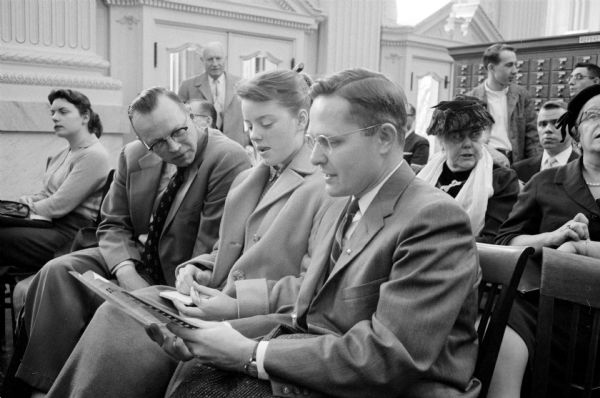 Three attendees at the symposium conducted by four national news commentators at the opening of the National Mass Communications History Center, held in the library of the Wisconsin State Historical Society. Left to right are Howard J. Rosenberry with his daughter, Peggy, and Rev. Erling Peterson, pastor of the Union Congregational Church in Windsor. Other people in the original image who were cropped out of the photograph published in the newspaper are not identified. A standing card catalog is seen in the background.