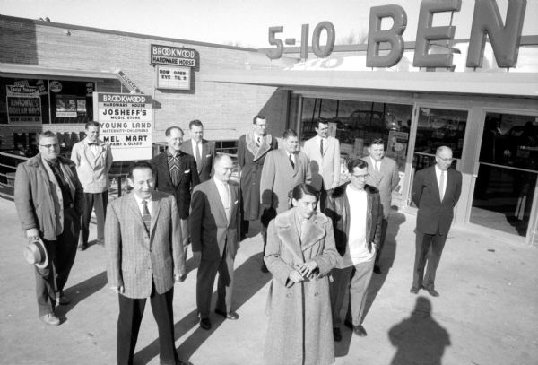 Elevated view of representatives of ten shops and two professional services gathered for the grand opening of the Brookwood Village shopping center at Nakoma Road and the West Beltline Highway. Part of the sign seen above them is for Ben Franklin's Five and Dime, and a marquee lists shops for Josheff's, Young Land and Mel Mart.