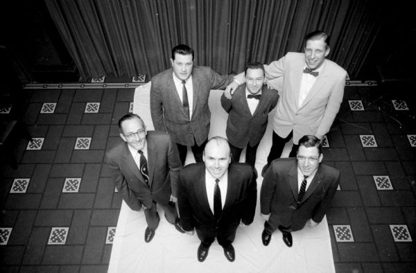 Overhead view of members of the Decem Associates, a Madison investment club, that claims it has the largest (by physical size) slate of officers in town. They are posing with the smallest member, 5' 11" Roy Morgenthaler in the center back row. Others are (front row, left to right): John Secord, club advisor; Joseph Fagen, president; Harold Scales, treasurer. In back are Richard Baer and Richard Stark, secretary. The camera angle is elevated with a downward perspective to show the tiled floor they are standing on.