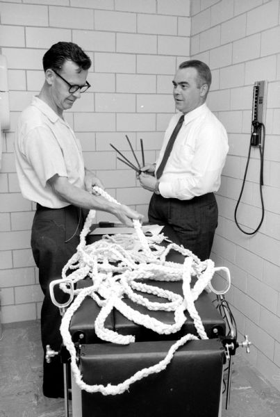 Two police detectives or officers, one handling hacksaw blades and the other inspecting rope, investigating a possible burglary using these items. They are standing in a tiled police station room.
