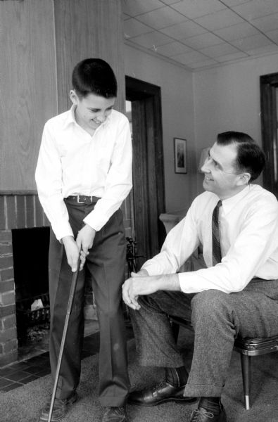 James Doyle, Jr. swinging a golf club under the gaze of his father, James Doyle, Sr. They are in a living room by the fireplace.