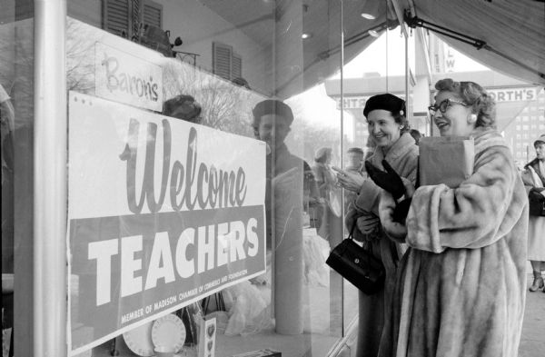 Teachers window shopping between sessions of the 66th Annual Southern Wisconsin Education Area convention. Left to right standing under the awning are  Eleanor Kolu and Ann Burwitz from Ithaca, WI. A poster in the front window for Baron's Department Store (at 12-18 West Mifflin Street) welcomes teachers.