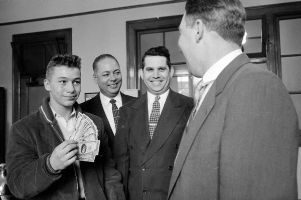 Warren Flisram (16, rural Brooklyn) is receiving $100 for his honesty in finding and helping recover $13,500 in cash along a railroad right-of-way on January 31, 1958. Warren found the money, most of it in $20 bills, after a mail pouch broke open as it was tossed out of a train and fell under the wheels. With Flisram are (left to right): John Cato, a postal inspector (Milwaukee); Donald Ferris, Brooklyn chief of police to whom Flisram reported his find; and Arthur Flisram, Warren's father.