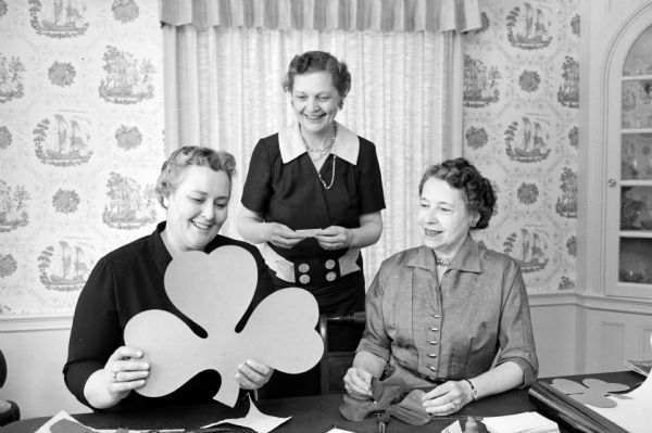 Three women at a table cutting paper shamrocks of different sizes. They are making the holiday crafts at a dining room table. 