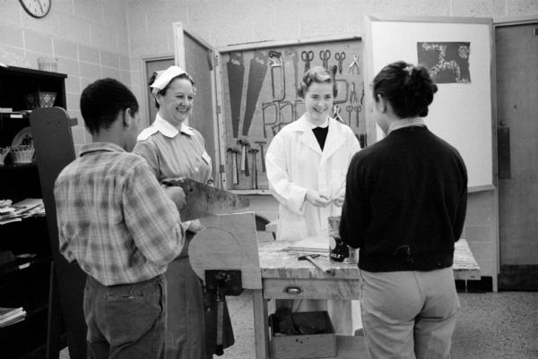 Red Cross Gray Ladies Mrs. Irving J. Maurer (left, 30 Fuller Drive) and Mrs. Ralph Penniall (right, 37-B University Houses) give advice and assistance to two patients in the occupational therapy unit of the Wisconsin Diagnostic Center at 1552 University Avenue. They are standing in front of a wall of hand tools.

The center was under the supervision of the Mental Hygiene Division of the State Department of Public Welfare and connected with the University of Wisconsin Medical School as a training ground for medical students and future psychiatrists.