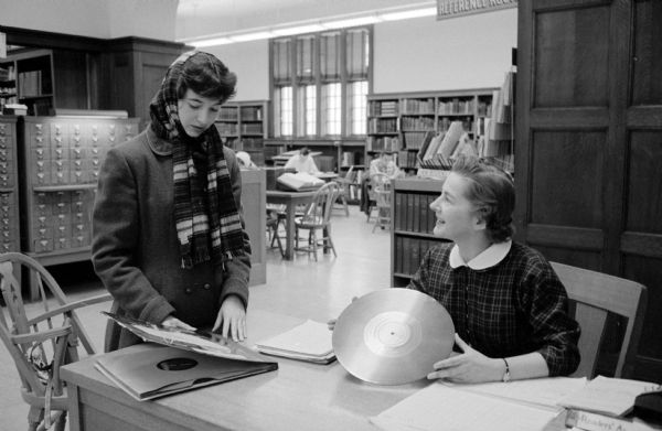 Peggy Van Wagenen (left, 122 E. Gilman Street) and Ellen Erickson (right, 1321 Morrison Street), check out an LP vinyl record in the Madison Central Library record library. Card catalogs and bookshelves are behind them.