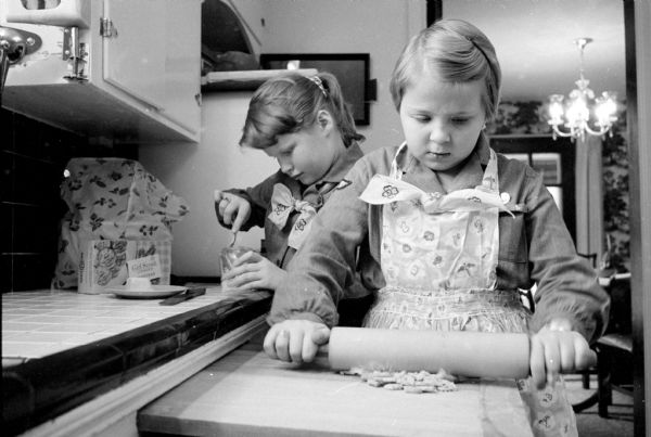 Two Madison Girl Scouts try out a pie recipe that has a crumb crust made with Girl Scout cookies. Elizabeth Davis (left) and Mary Beth Gehner are shown at work in a kitchen preparing the crust. Mary Beth, wearing an apron, is using a rolling pin to create the cookie crumble.