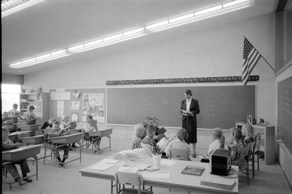 Scene of a second-grade classroom with students and their teacher, Helen G. Smith, who is standing in front of the blackboard. The photograph was taken to highlight the modern features of classrooms, like lighting and long chalkboards, as compared to classrooms of the 1920's.
