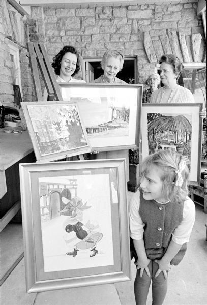 Garden Club floral paintings. Four paintings displayed with three women standing behind paintings and one young girl standing in front looking at a scene of a goose dressed in a floppy hat, dress and boots. The building is made of cut rock. 