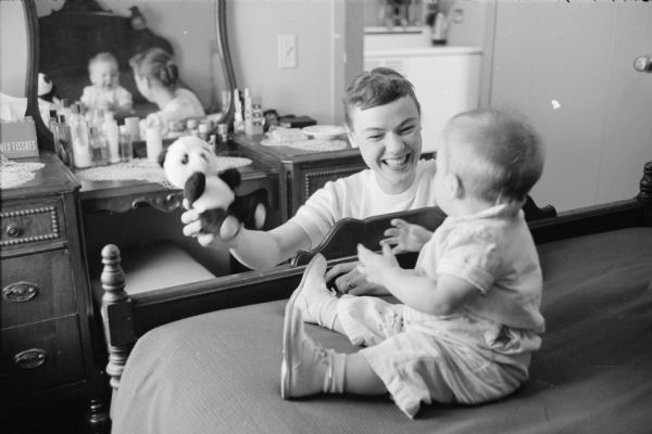 Mrs. Paul A. Weinhold and 8-month-old daughter, Julie, playing in their U.W. Eagle Heights apartment. Mr. Weinhold, a graduate of Colorado State University at Fort Collins, is studying for his masters degree in biochemistry. The apartment rents for $72 per month. Mrs. Weinhold is seen holding a stuffed panda bear as Julie is sitting at the end of the bed.    