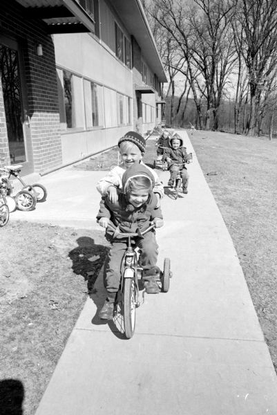 Children enjoy riding bikes at the U.W. Eagle Heights apartments. Barbara Bessae, daughter of Mr. and Mrs. Frank Bessae, is shown giving a ride to Tommy Biwer (4), son of Mr. and Mrs. Richard Biwar. Two unidentified bike riders are in the background on the sidewalk.