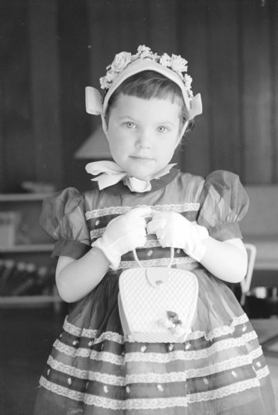 3-year-old Betsy Cashin, daughter of Mr. and Mrs. Robert Cashin, is shown in dress and gloves, wearing her Easter bonnet and holding a matching purse in front of her.