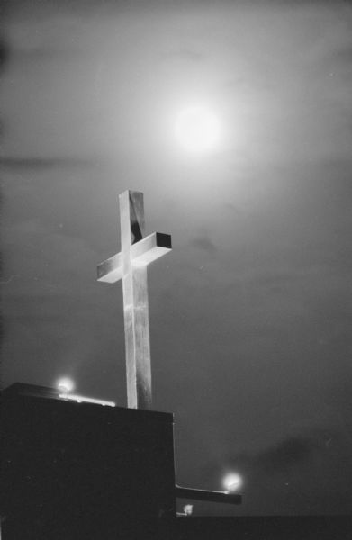 A cross at night at top of unidentified Christian church. The cross is illuminated by lamps with the moon shining through the clouds behind.