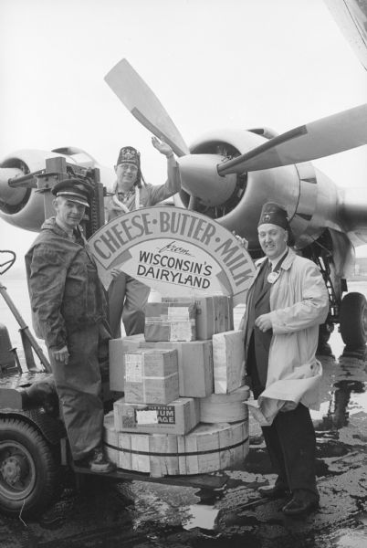 Rufus Wells, back, and Leo W. Roethe, right, standing beside the 350 pounds of cheese the Madison Zor Shrine group took with them on their tour of Hawaii. A man wearing work clothes and a hat is on the left, standing on the fork lift truck on which the cheese is stacked. An airplane is parked behind the men.