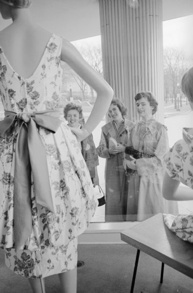 Three women standing outdoors on a sidewalk viewing a dress on a mannequin in the foreground inside a show window on Capitol Square. They are (L-R): Linda Schereck; Shelagh Reid, president of the Winnipeg Y-Teens, and Joan Clifford. The Wisconsin State Capitol is in the background across the street.