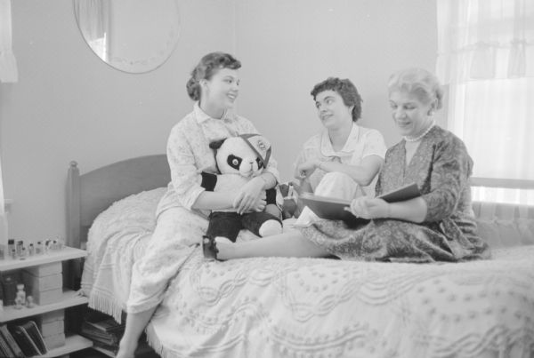 "All the Winnipeg girls were housed with Madison families during their four-day stay. While Bonnie Rejahl, hugging a toy bear, left, and her Canadian house guest, Valdeen Bronwell, center, indulge in some teen-aged chatter before bedtime, Bonnie's mother, Mrs. C.D. Rejahl, thumbed through Valdeen's high school yearbook." The group is sitting on top of the bed.