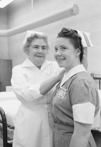 One of the philanthropic projects of the Madison Altrusa Club's annual country fair proceeds is scholarships for young women to take the practical nurse's course at the Madison Vocational school. Ruth Cole, registered nurse and director of the course places a cap on Beverly Brown, one of the new practical nurses. The fair is held on the Spruce Hill farm near Cambridge.