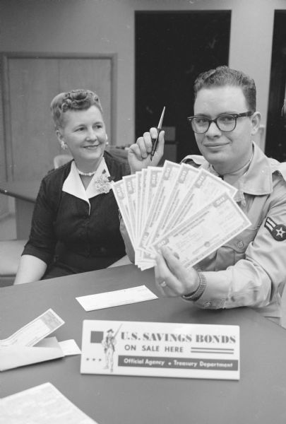 "Airman 2/C Don R. Roberts, Truax Field, is shown purchasing his portion of 'Share in America' — 15 U.S. $50 savings bonds. Roberts, a native of Wichita Falls, Texas, is shown with Clerk Hazel Micka of the Bank of Madison."  