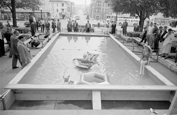 Model Marlyn Sachtjen paddling around in the 16 by 32 foot pool which was constructed on the Capitol Park overlooking State Street. Ruth Andres sits at the edge of the pool modeling sportswear as part of the display. The "Outdoor Living Day" event is sponsored by the Madison Chamber of Commerce. 